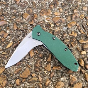 Puede incluir: Una navaja de bolsillo plegable verde con una hoja plateada. La navaja est&aacute; abierta, mostrando el filo de la hoja. El mango es de color verde liso y est&aacute; sobre una superficie de piedra texturizada.