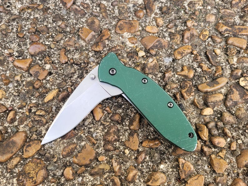 Puede incluir: Una navaja de bolsillo plegable verde con una hoja plateada. La navaja est&aacute; abierta, mostrando el filo de la hoja. El mango es de color verde liso y est&aacute; sobre una superficie de piedra texturizada.