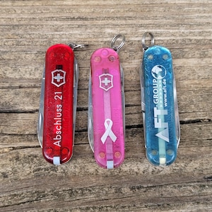 Può includere: Tre coltelli svizzeri colorati su una superficie di legno. I coltelli sono rossi, rosa e blu. Il coltello rosso ha il testo "Abschluss 21". Il coltello rosa ha un simbolo a nastro bianco. Il coltello blu ha il testo "AFT-GROUP".