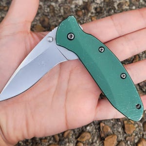 Puede incluir: Una navaja plegable con una hoja plateada y un mango verde. La hoja est&aacute; parcialmente abierta, revelando su filo. El mango tiene tres remaches negros y un peque&ntilde;o agujero en el extremo. La navaja est&aacute; dise&ntilde;ada para el uso diario.