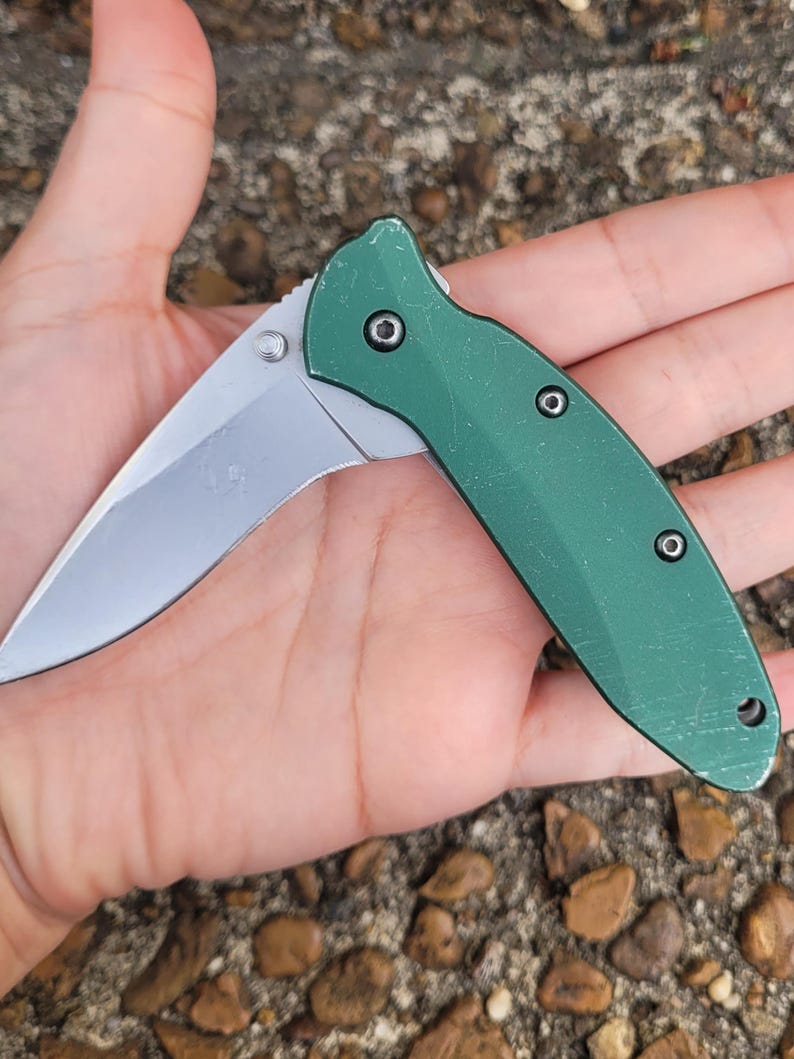 Puede incluir: Una navaja plegable con una hoja plateada y un mango verde. La hoja est&aacute; parcialmente abierta, revelando su filo. El mango tiene tres remaches negros y un peque&ntilde;o agujero en el extremo. La navaja est&aacute; dise&ntilde;ada para el uso diario.