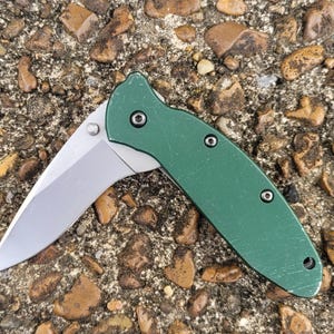 Puede incluir: Un cuchillo de bolsillo plegable verde con una hoja plateada est&aacute; abierto, mostrando su filo. El mango tiene una superficie texturizada y est&aacute; asegurado con tres tornillos peque&ntilde;os. El cuchillo descansa sobre un lecho de peque&ntilde;as rocas.