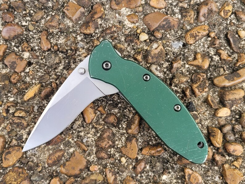 Puede incluir: Un cuchillo de bolsillo plegable verde con una hoja plateada est&aacute; abierto, mostrando su filo. El mango tiene una superficie texturizada y est&aacute; asegurado con tres tornillos peque&ntilde;os. El cuchillo descansa sobre un lecho de peque&ntilde;as rocas.