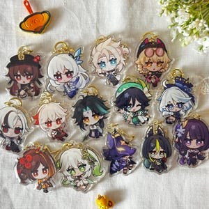 Puede incluir: Una colección de llaveros acrílicos con diseños de personajes coloridos de estilo anime. Cada llavero tiene un anillo dorado para sujetar. Los personajes tienen varios colores de pelo y atuendos, con expresiones faciales detalladas.