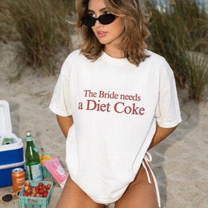 Pode incluir: Camiseta creme com o texto vermelho "The Bride needs a Diet Coke". A cena mostra uma praia com um cooler, bebidas e lanches. A pessoa está usando óculos de sol.