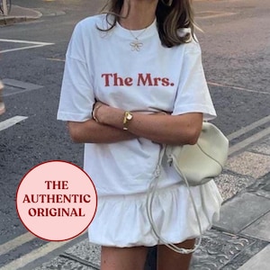 Könnte beinhalten: Eine Frau trägt ein weißes T-Shirt-Kleid mit dem roten Text "The Mrs.". Sie trägt rote Sandalen mit goldenen Schnallen und eine weiße Tasche. Das Bild zeigt eine Straßenszene.