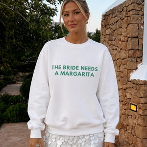 May include: A white crewneck sweatshirt featuring the green text THE BRIDE NEEDS A MARGARITA printed on the chest, paired with a short white skirt covered in circular sequins and a white quilted handbag with a knotted handle design.
