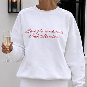 May include: A white long-sleeve crewneck sweatshirt featuring the red cursive text If lost please return to Nick Morrison on the chest, paired with a matching white mini skirt that has a small vertical slit on the front hemline.