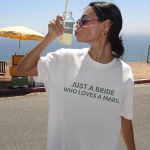 May include: A woman wearing a white t-shirt with the text "JUST A BRIDE WHO LOVES A MARG." printed on it. She is holding a glass bottle with a straw and is looking at the camera.