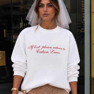 May include: A white crewneck sweatshirt featuring the red embroidered text If lost please return to Calum Lewis, paired with a sheer white bridal veil and a gold sequined skirt. The sweatshirt has long sleeves and a relaxed fit, creating a casual yet formal look.