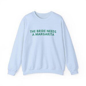 May include: A light blue crewneck sweatshirt featuring the text THE BRIDE NEEDS A MARGARITA printed in dark green capital letters across the chest. The garment has long sleeves, a ribbed neckline, and ribbed cuffs at the wrists and the bottom hem.