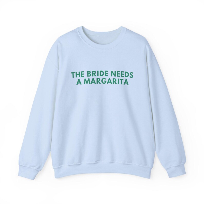 May include: A light blue crewneck sweatshirt featuring the text THE BRIDE NEEDS A MARGARITA printed in dark green capital letters across the chest. The garment has long sleeves, a ribbed neckline, and ribbed cuffs at the wrists and the bottom hem.