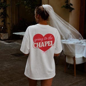 May include: A white short-sleeved t-shirt featuring a large red heart graphic on the back with the text "going to the CHAPEL" printed in white and a small pink bow illustration at the bottom of the heart, paired with a sheer white veil.