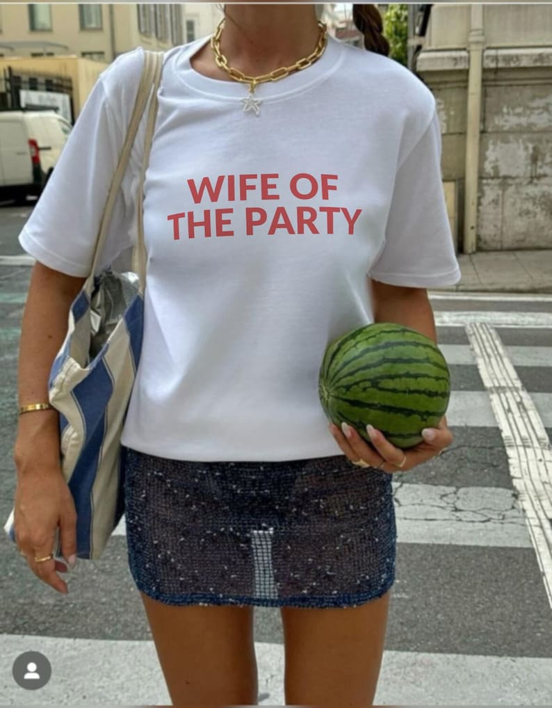 May include: White t-shirt with the text "WIFE OF THE PARTY" in red. The person is wearing a gold necklace and a sequined mini skirt. A striped tote bag is over the shoulder. A small watermelon is held in the hand.