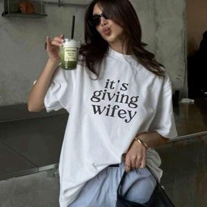 May include: A woman wearing a white t-shirt with the text "it's giving wifey" in black lettering. She is wearing blue jeans and a black handbag. She is holding a green drink in her hand.