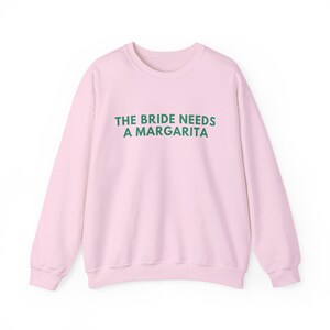 May include: A light pink long-sleeve crewneck sweatshirt featuring the text THE BRIDE NEEDS A MARGARITA printed in dark green capital letters across the chest. The garment has a ribbed neckline, cuffs, and hem, with a solid color design.
