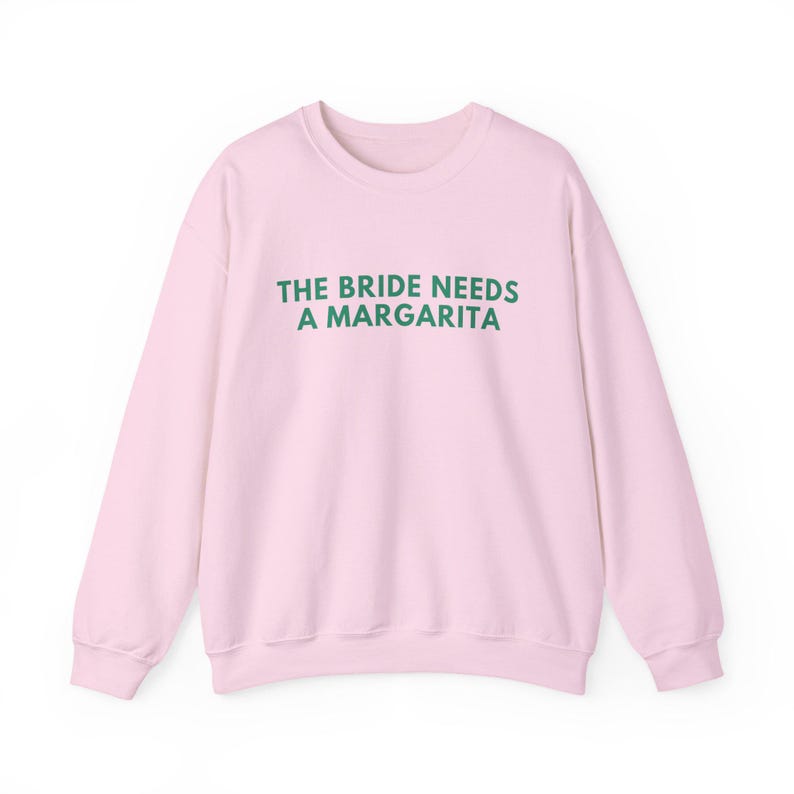 May include: A light pink long-sleeve crewneck sweatshirt featuring the text THE BRIDE NEEDS A MARGARITA printed in dark green capital letters across the chest. The garment has a ribbed neckline, cuffs, and hem, with a solid color design.