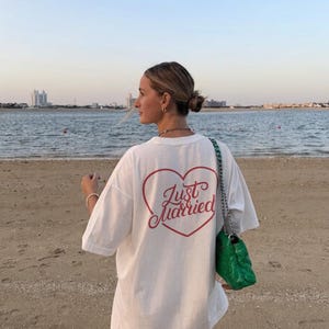 May include: White oversized t-shirt with a red heart-shaped graphic and the words "Just Married" in a script font. The shirt is worn on a beach with a green handbag.