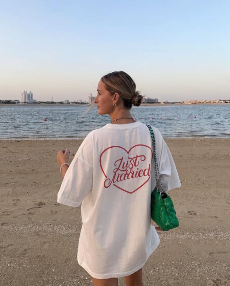 May include: White oversized t-shirt with a red heart-shaped graphic and the words "Just Married" in a script font. The shirt is worn on a beach with a green handbag.