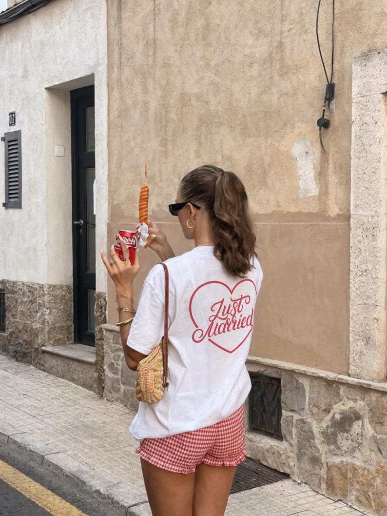May include: A white t-shirt with a red heart-shaped graphic that says "Just Married" on the back. The person is wearing red and white checkered shorts and holding a red can. A small woven bag hangs from the shoulder.
