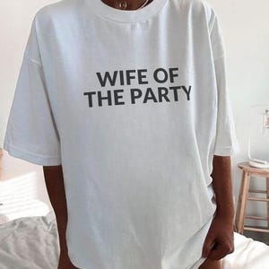 May include: White t-shirt with the text "WIFE OF THE PARTY" in black block letters. The shirt has short sleeves and a crew neck. The shirt is worn casually.