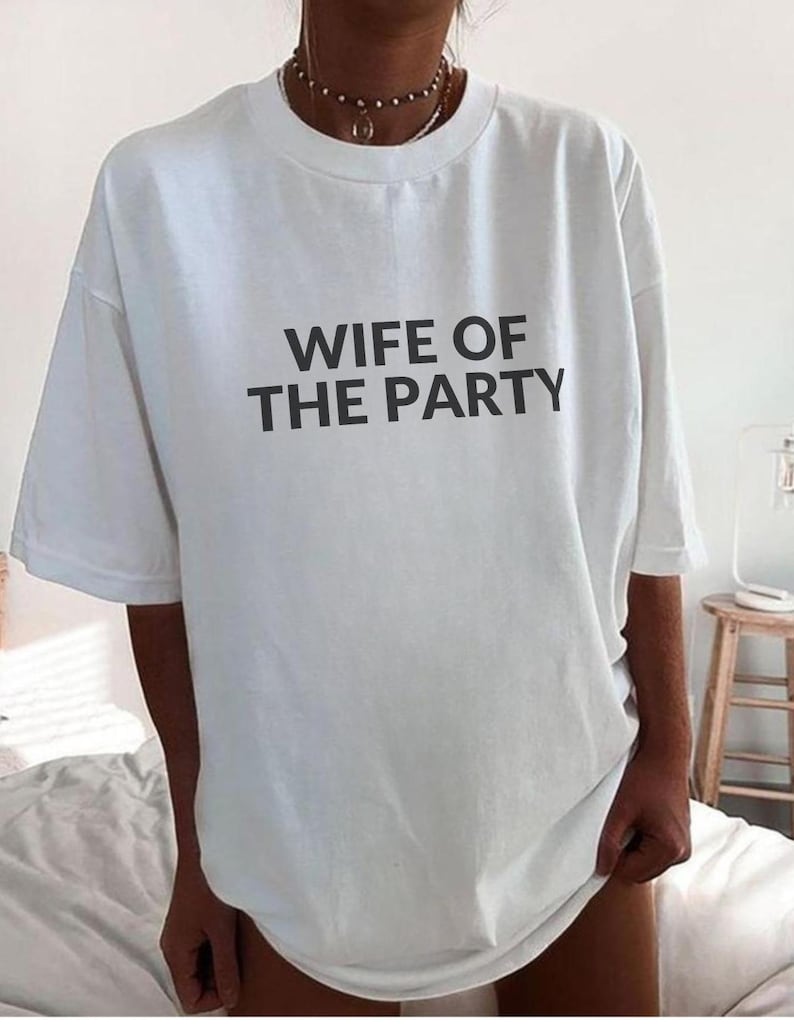 May include: White t-shirt with the text "WIFE OF THE PARTY" in black block letters. The shirt has short sleeves and a crew neck. The shirt is worn casually.