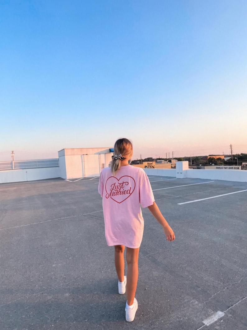May include: A light pink t-shirt with the words "Just Married" inside a heart graphic on the back. The shirt is worn with white sneakers. The background is a rooftop parking lot with a blue sky.