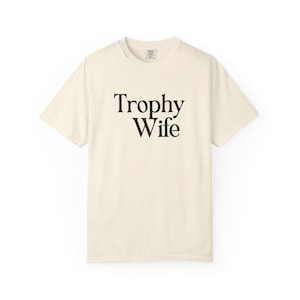 May include: Cream-colored t-shirt with the words "Trophy Wife" printed in black, centered on the front. The shirt has a classic crew neck and short sleeves, made of a soft, comfortable material.
