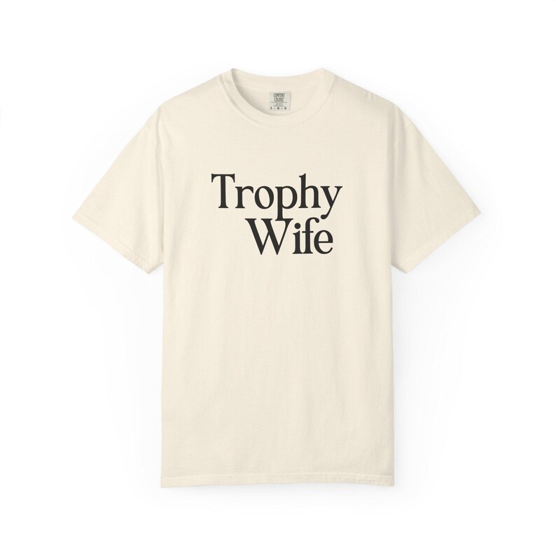 May include: Cream-colored t-shirt with the words "Trophy Wife" printed in black, centered on the front. The shirt has a classic crew neck and short sleeves, made of a soft, comfortable material.