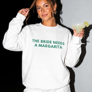 May include: A white crewneck sweatshirt featuring the dark green text THE BRIDE NEEDS A MARGARITA printed on the chest, paired with white lace shorts. The garment is worn with a glass containing a margarita garnished with a lime slice.