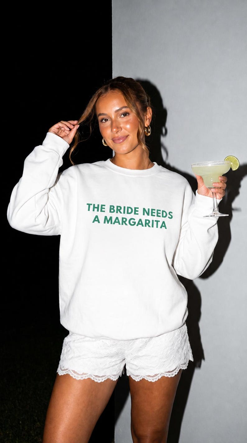 May include: A white crewneck sweatshirt featuring the dark green text THE BRIDE NEEDS A MARGARITA printed on the chest, paired with white lace shorts. The garment is worn with a glass containing a margarita garnished with a lime slice.