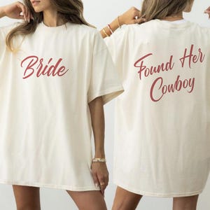 Könnte beinhalten: Cremefarbene T-Shirts mit bordeauxrotem Text. Eines der Shirts trägt vorne den Schriftzug "Bride". Das andere Shirt trägt hinten "Found Her Cowboy". Die Shirts haben kurze Ärmel und eine lockere Passform.