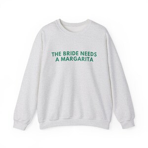 May include: A light gray heathered crewneck sweatshirt featuring the text THE BRIDE NEEDS A MARGARITA printed in dark green capital letters across the chest. The garment has long sleeves, a ribbed neckline, and ribbed cuffs at the wrists and hem.