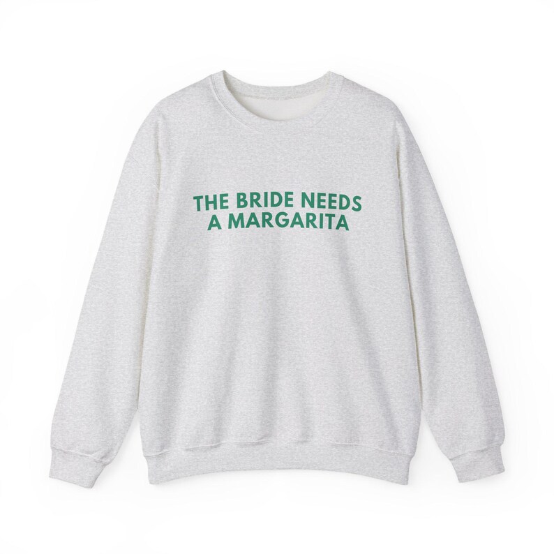 May include: A light gray heathered crewneck sweatshirt featuring the text THE BRIDE NEEDS A MARGARITA printed in dark green capital letters across the chest. The garment has long sleeves, a ribbed neckline, and ribbed cuffs at the wrists and hem.