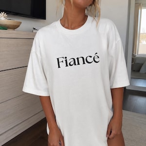 May include: A white short-sleeved t-shirt with a crew neckline featuring the word Fiancé printed in a black serif font across the center of the chest. The garment has a loose, relaxed fit with dropped shoulders and a straight hemline.