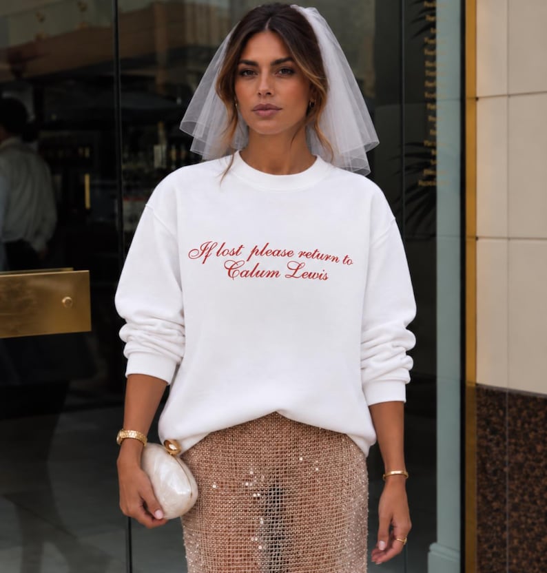 May include: A white crewneck sweatshirt featuring the red embroidered text If lost please return to Calum Lewis, paired with a sheer white bridal veil and a gold sequined skirt. The sweatshirt has long sleeves and a relaxed fit, creating a casual yet formal look.