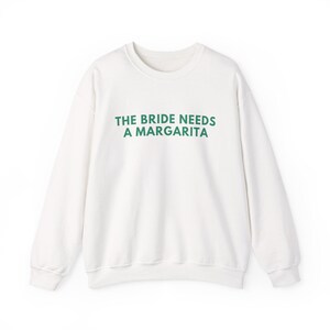 May include: A white long-sleeve crewneck sweatshirt featuring the text THE BRIDE NEEDS A MARGARITA printed in dark green capital letters across the chest. The garment has a ribbed neckline, cuffs, and hem, with a simple, clean design.