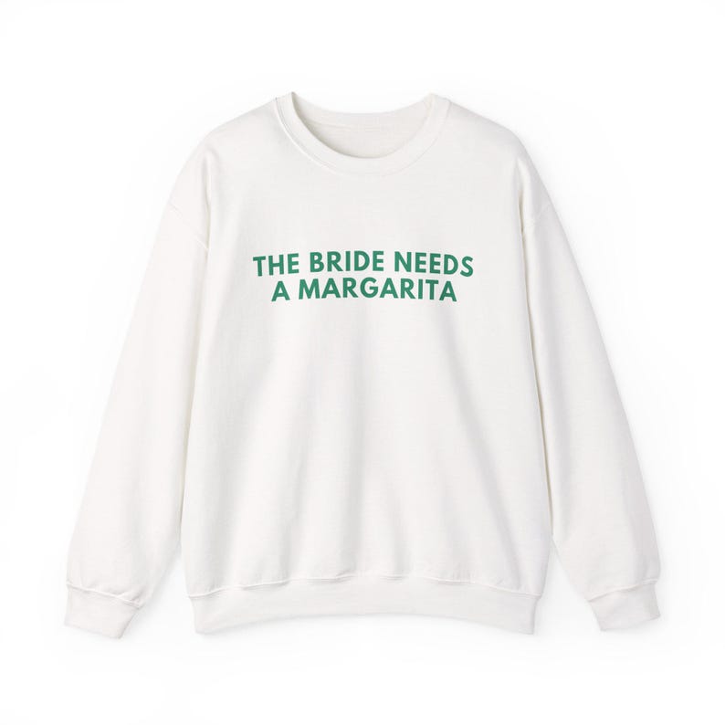 May include: A white long-sleeve crewneck sweatshirt featuring the text THE BRIDE NEEDS A MARGARITA printed in dark green capital letters across the chest. The garment has a ribbed neckline, cuffs, and hem, with a simple, clean design.