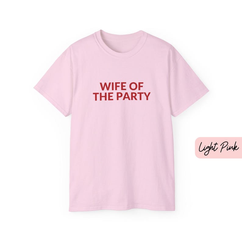 May include: Light pink t-shirt with the text "WIFE OF THE PARTY" in red. The shirt has short sleeves and a crew neck. A small label in the bottom right corner reads "Light Pink".