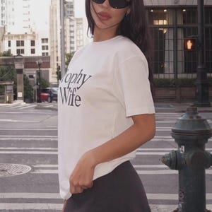 May include: A woman wearing a white t-shirt with the words "Trophy Wife" in black lettering, paired with a dark gray skirt and black sunglasses. The background shows a city street with buildings and a fire hydrant.