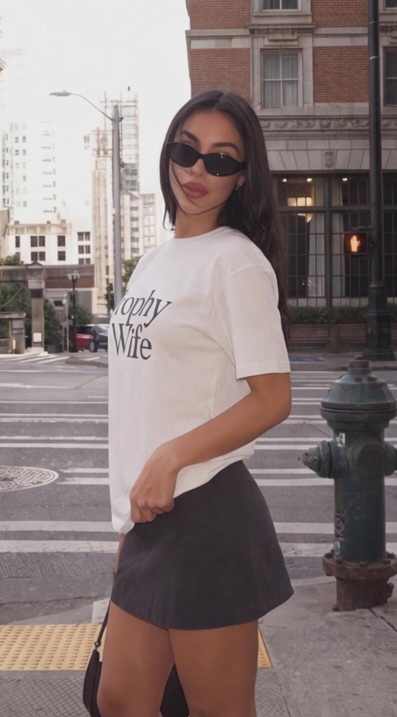 May include: A woman wearing a white t-shirt with the words "Trophy Wife" in black lettering, paired with a dark gray skirt and black sunglasses. The background shows a city street with buildings and a fire hydrant.