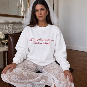 May include: A white long-sleeve crewneck sweatshirt featuring red cursive text that reads If lost please return to Robert Mills, paired with a white sheer lace pant with a floral pattern and a white tulle bridal veil worn on the head.