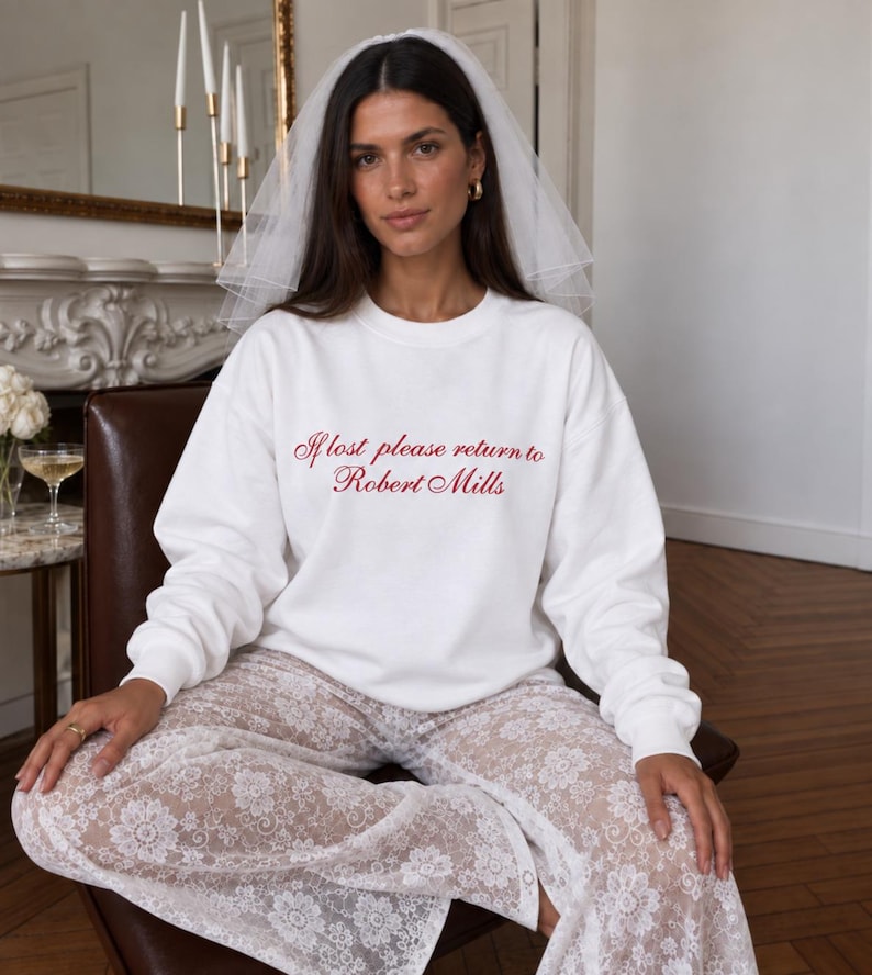 May include: A white long-sleeve crewneck sweatshirt featuring red cursive text that reads If lost please return to Robert Mills, paired with a white sheer lace pant with a floral pattern and a white tulle bridal veil worn on the head.