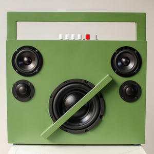 Pode incluir: Um alto-falante verde retangular com uma alça. Possui cinco alto-falantes pretos de vários tamanhos, botões brancos e vermelhos e uma barra verde diagonal. O design lembra um boombox.