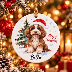 Op de afbeelding: Een ronde witte keramische kersthanger met een aquarelillustratie van een bruin-witte puppy met een rode kerstmuts. Een groene kerstboom, blauwe sneeuwvlokken, de tekst Merry Christmas en de naam Bella zijn op het ornament gedrukt.