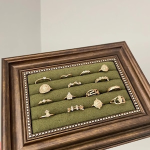 Pode incluir: Uma coleção de anéis dourados exibidos em uma caixa de joias com moldura de madeira e interior de veludo verde. Os anéis apresentam vários designs, incluindo padrões florais, geométricos e texturizados. A moldura é marrom escuro.