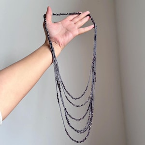 Puede incluir: Un collar de múltiples hilos con cuentas de color púrpura oscuro. El collar se sostiene sobre un fondo blanco liso. Los hilos caen en cascada en un efecto de capas, creando un aspecto sofisticado y elegante. Las cuentas parecen pequeñas y facetadas, captando la luz.