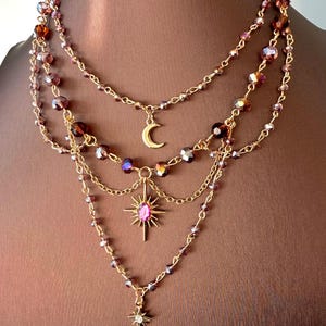 Könnte beinhalten: Mehrreihige Halskette mit goldenen Ketten und weinroten Perlen. Sie hat einen Halbmond, einen Sternen-Anhänger mit rosa Mitte und einen kleinen Stern-Anhänger. Die Halskette wird vor einem braunen Hintergrund präsentiert.