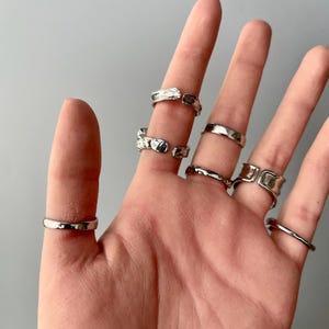 May include: A collection of silver rings in various designs. The rings are worn on multiple fingers, showcasing different styles, including smooth bands, textured surfaces, and open-ended designs. The rings are displayed against a neutral gray background.