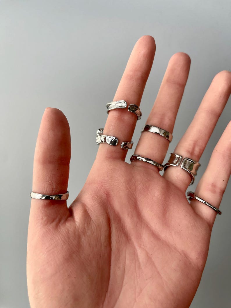 May include: A collection of silver rings in various designs. The rings are worn on multiple fingers, showcasing different styles, including smooth bands, textured surfaces, and open-ended designs. The rings are displayed against a neutral gray background.
